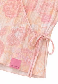 Κοντινή λήψη ενός ροζ και ροδακινί floral wrap τοπ με δέσιμο στο πλάι και ροζ ετικέτα με το γράμμα OH APRIL στο τελείωμα.