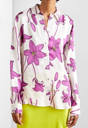 Weiße Bluse mit Knopfleiste und großem rosa Blumenmuster, getragen über einem knallgelben Rock bei einer stehenden Person.