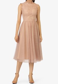 Robe sans manches en dentelle rose poussiéreux avec col montant et jupe midi en tulle, portée avec des talons transparents à lanière au niveau de la cheville.