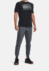 Svart t-shirt med kort ärm med "UNDER ARMOUR" grafik i silver; grå tapered byxor med en slät yta; svarta träningsskor med orange detaljer.