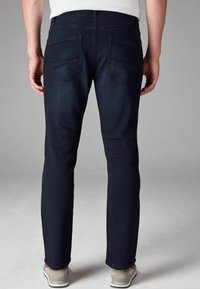 Los jeans de mezclilla oscura cuentan con un corte slim, una cintura de media altura y dos bolsillos traseros con detalles de costura. La tela tiene una textura suave y ligeramente desgastada.