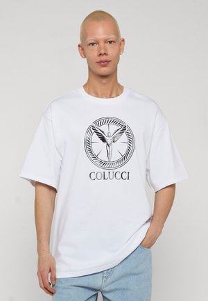 Ung person med kort blondt hår iført hvid oversize T-shirt med sort vinget figur og teksten "COLUCCI", kombineret med lyseblå jeans.