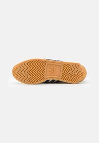 Zapatilla con una suela de goma beige que presenta un patrón de tracción en espiga. Parte superior de lona azul marino con tres rayas blancas a lo largo del costado.
