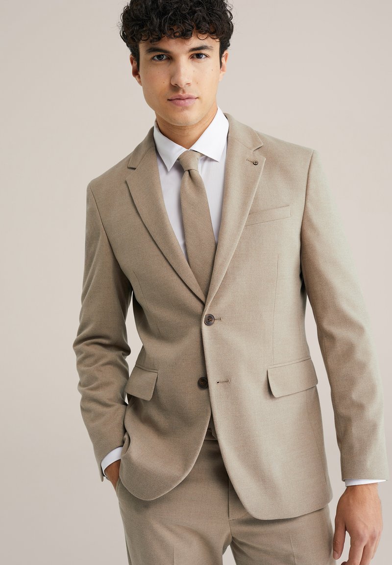 Beige színű, finom anyagból készült szabott háromrészes öltöny; jellegzetes hajtókával, két gombbal és illő nyakkendővel rendelkezik.