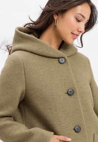 Junge Frau mit langen dunklen Haaren, die silberne Creolen und einen taupefarbenen Wollmantel mit großen schwarzen Knöpfen und einem breiten Kragen trägt.