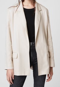 Světlý béžový blazer vyrobený z hladké tkaniny, s strukturovaným designem, výstřihy do tvaru notche a dvěma předními náprsními kapsami. Nositý přes černý top.