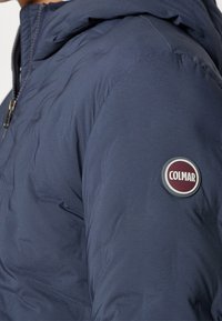 Donkerblauwe gewatteerde jas met mouw en een ronde 'Colmar' merklogopatch en een gedeeltelijk zichtbare ritssluiting aan de voorkant.