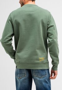 Groene sweatshirt met een ronde hals, gestructureerde stof en gele geborduurde details aan de linkerbenedenkant. Gepaard met blauwe jeans.