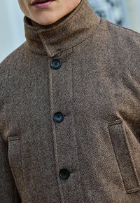Ruskea herringbone-kuvioinen takki, jossa on korkea kaulus, kolmonen mustaa nappikiinnitystä ja kaksi etutaskua. Teksturoitu kangas näkyvissä.