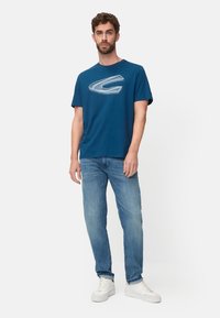 Men's blue cotton t-shirt featuring a large white graphic logo, paired with light blue denim jeans and white sneakers. Simple, casual design.