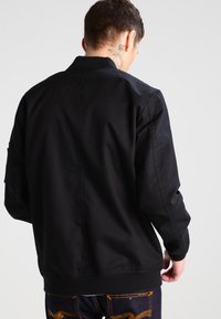 Juodas bomber tipo švarkas su lygia audinio tekstūra, megztu apatiniu kraštu ir rankogaliais; pasižymi minimalistiniu dizainu be matomų logotipų ar raštų.