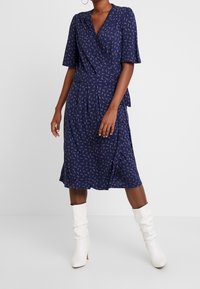 Kobieta w granatowej sukience w kwiaty z rękawami do łokci i białych, sięgających do połowy łydki szpilkach, na jednolitym tle.