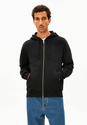 Mann mit lockigem Haar und Schnurrbart, der einen schwarzen Hoodie mit Reißverschluss trägt, Hände in den Taschen und blaue Jeans vor weißem Hintergrund.