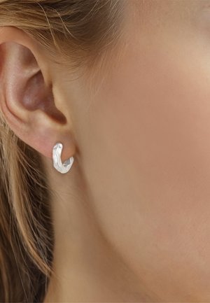 Gros plan d'une oreille à la peau claire portant une petite boucle d'oreille en argent martelé avec une texture ondulée.