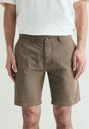 Bărbat purtând pantaloni scurți maro până la genunchi și un tricou alb, în picioare cu brațele relaxate pe lângă corp, pe un fundal simplu.