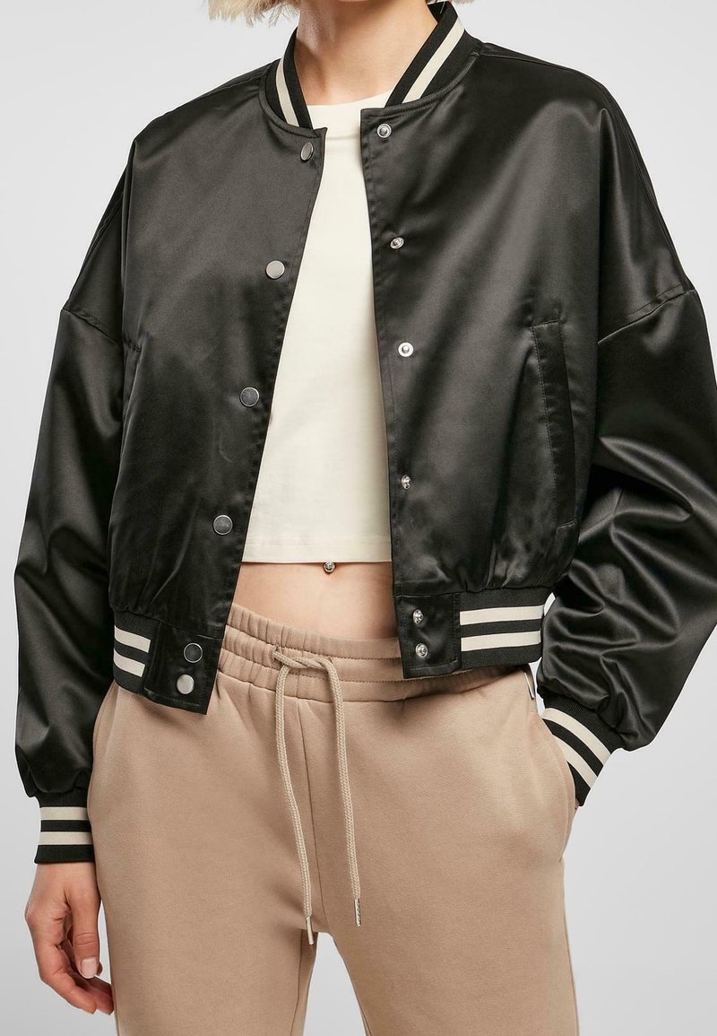 Personne portant une veste bomber en satin noire avec poignets et bas rayés, un top blanc court et un pantalon beige à cordon, les mains dans les poches.