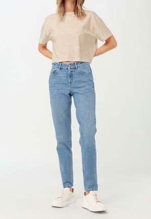 Persoon die een beige cropped t-shirt, lichtblauwe high-waisted jeans en witte platformsneakers draagt, staande met de handen achter de rug.