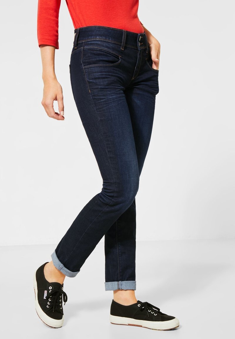 Jean bleu foncé slim-fit retroussé aux chevilles, porté avec des baskets noires et un haut rouge à manches longues, debout devant un fond clair uni.