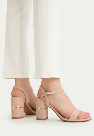 Pies con sandalias beige de tacón cuadrado y punta abierta, con correas en el tobillo, combinadas con pantalones blancos de pierna ancha sobre un fondo claro.