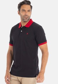 Homme portant un polo noir avec un col et des bords de manches rouges, un pantalon beige, se tenant devant un fond clair uniforme.