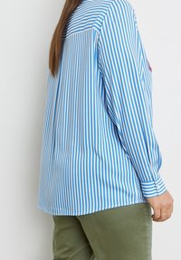 Blue and white striped long-sleeve shirt featuring a button cuff and back pleat, paired with olive green pants. Smooth texture.