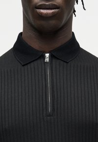 Gros plan d'une personne portant une chemise noire côtelée avec un col texturé et une fermeture éclair argentée partiellement fermée au niveau du cou.