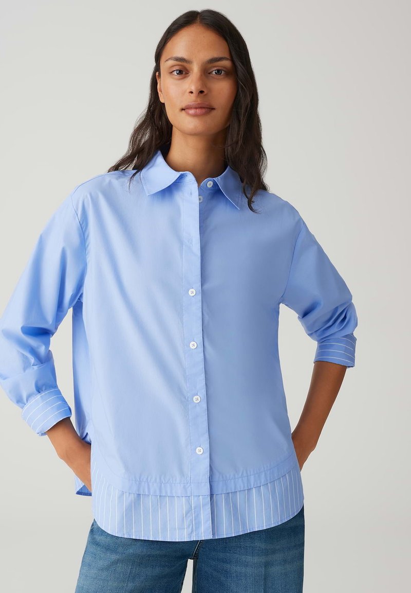 Woman with long dark hair wears a light blue button-up shirt with striped cuffs and hem, paired with blue jeans on a plain background.