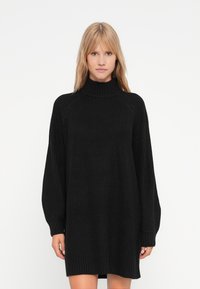 Melna adīta kleita ar augstu apkakli, garām piedurknēm un ribotu aproci. Iespējo brīvu piegriezumu un strukturētu rakstu. Gluda auduma.