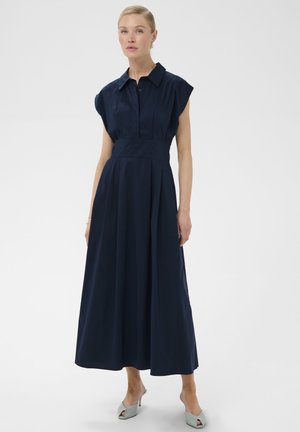 Femme portant une robe midi sans manches bleu marine avec un col et une jupe plissée, associée à des mules à talons ouvertes gris clair.