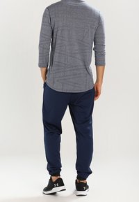 Man som står med ryggen vänd, iförd en grå långärmad skjorta, marinblå joggingbyxor och svarta Under Armour-sneakers.