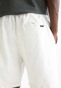 Shorts en coton blanc avec une ceinture élastique, dotés d'une poche arrière unique et d'une petite étiquette avec logo noir près de la poche.