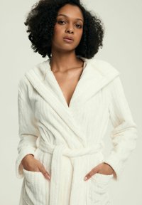Kobieta o kręconych włosach, ubrana w biały pluszowy szlafrok z kapturem i kieszeniami, z rękami spoczywającymi w kieszeniach na jednolitym tle.