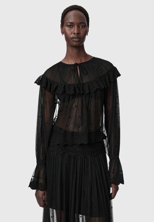 Femme portant un chemisier noir transparent avec des détails de volants et de dentelle, manches longues et col noué, sur un fond gris uni.