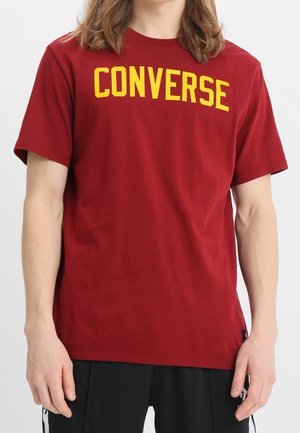 T-shirt en coton rouge avec un grand logo jaune "CONVERSE" incurvé sur la poitrine. Col rond classique et manches courtes.