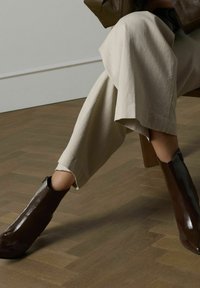 Bruine enkellaarsjes van glanzend leer, met een scherp silhouet en een ritssluiting. Draag ze met lichtbeige wijde broek op een houten vloer.