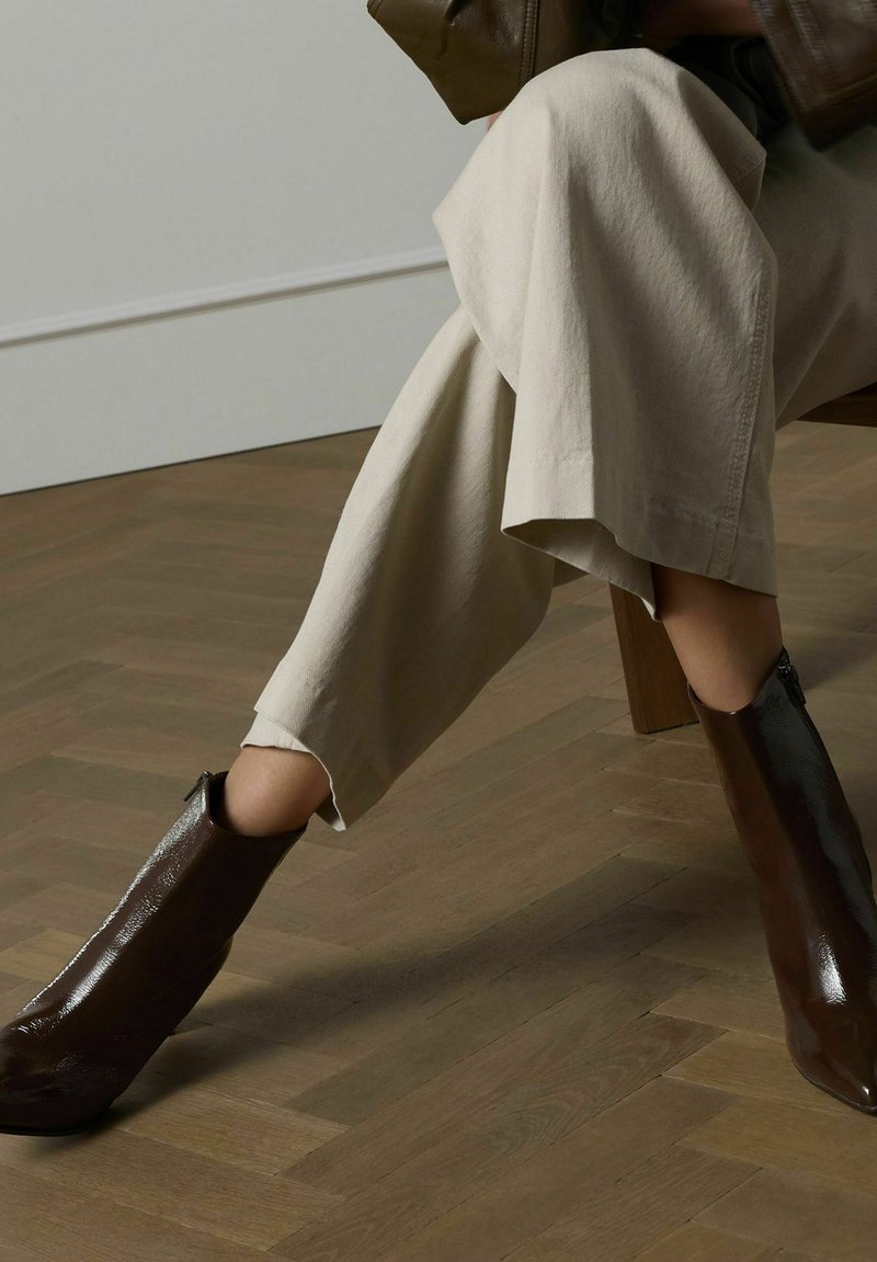 Bruine enkellaarsjes van glanzend leer, met een scherp silhouet en een ritssluiting. Draag ze met lichtbeige wijde broek op een houten vloer.