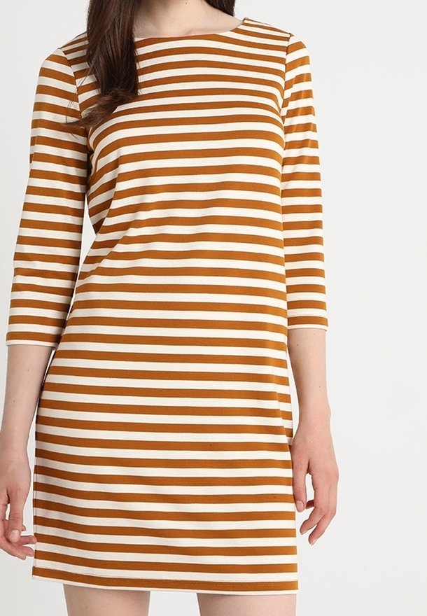 Femme portant une robe rayée horizontalement marron et blanche avec des manches trois-quarts, debout devant un fond uni.