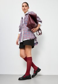 Geruite gelaagde shirt in paars en rood, zwarte shorts, bordeaux kniehoge sokken en zwarte loafers met een gesp. Draagt een bordeaux tas.