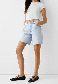 Kvinna som bär ljusblå denimknä-långa shorts, vit kortärmad topp och svarta mesh Mary Jane-sandaler, stående med ena handen i fickan.