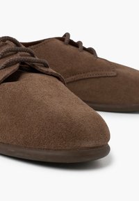 Scarpe in suede marrone con punta arrotondata, caratterizzate da un design semplice, lacci tonali e una suola in gomma liscia. La texture sembra morbida e opaca.