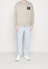 Felpa beige con logo "AX" nero sul petto, abbinata a jeans dritti blu chiaro e sneakers bianche. Tessuto morbido, vestibilità comoda.