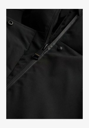 Zwarte jasstof met een gladde textuur, voorzien van een ritsclosure en een ronde drukknoop. De rits trekt wordt aangeduid met "SUPERDRY."