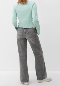 Mátově zelený žebrovaný top s dlouhým rukávem a vlnitým lemem, v kombinaci s šedými džíny s vysokým pasem a širokými nohavicemi, které mají dvě zadní kapsy a švy.