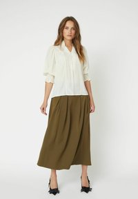 Bílá blůza s nabíranými rukávy, s detailními knoflíky, a plisovaná olivová sukně. Černé boty s mašličkovými akcenty dokončují outfit.