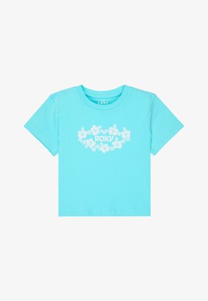 Mädchen mit langen Haaren, das ein hellblaues kurzes T-Shirt mit floralem Muster und dunkelblaue Jeansshorts trägt, posiert mit einer Hand auf der Hüfte.