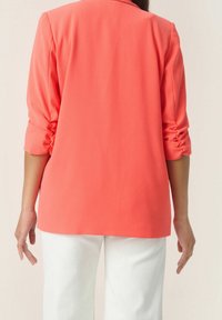 Koralowy żakiet z marszczonymi rękawami, gładka tkanina; połączony z białymi, szerokimi spodniami. Widok od tyłu ukazuje dostosowaną sylwetkę i czyste linie.