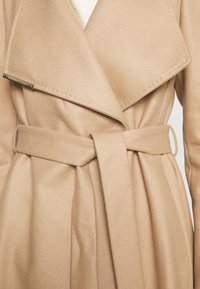 Manteau en laine beige avec larges revers et ceinture en tissu nouée à la taille, porté sur un haut blanc côtelé.