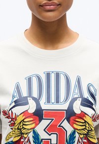 Άτομο που φοράει λευκό μπλουζάκι με μπλε κείμενο "ADIDAS", μεγάλο κόκκινο αριθμό 3 και δύο πολύχρωμους τούκους που κοιτάζουν ο ένας τον άλλον.