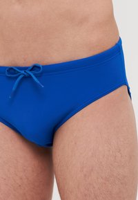 Modre kopalne hlače iz gladkega materiala, z vrvico za zavezovanje, nizkim krojem in minimalnimi stranskimi odprtinami.