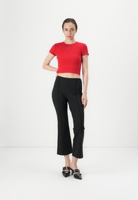 Raudona trumpa marškinė su mėlynu logotipu, derinamos su juodomis platėjančiomis kelnėmis. Modelis nešioja juodas aukštakulnes basutes su apvaliais blizgučiais. Minimalistinis fonas.